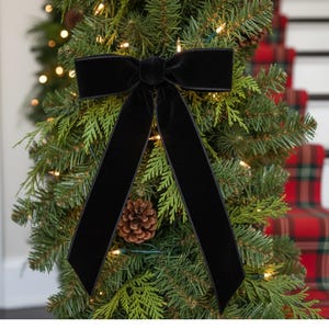 May include: A black velvet bow adorns a Christmas tree, its long tails cascading over the evergreen branches. A pine cone rests among the needles, and warm white lights twinkle, creating a festive holiday scene.