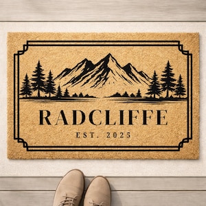 May include: A rectangular doormat with a mountain and tree silhouette design. The word "RADCLIFFE" is displayed in a bold font, with "EST. 2025" below. The mat is tan with a black border and design.