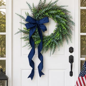 May include: A green eucalyptus wreath with a navy blue bow and streamers hangs on a white door. The wreath is composed of various shades of green foliage. A black door handle and lock are visible.