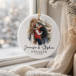 K&ouml;nnte beinhalten: Runder wei&szlig;er Keramikschmuck mit goldenem Aufh&auml;ngeband. Er zeigt ein Aquarellbild eines sich k&uuml;ssenden Paares. Die Namen "Jennifer & Stephen" und das Wort "ENGAGED" mit dem Datum "2.14.2026" sind in schwarzer Schrift geschrieben.