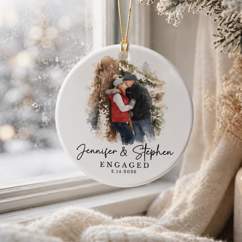 K&ouml;nnte beinhalten: Runder wei&szlig;er Keramikschmuck mit goldenem Aufh&auml;ngeband. Er zeigt ein Aquarellbild eines sich k&uuml;ssenden Paares. Die Namen "Jennifer & Stephen" und das Wort "ENGAGED" mit dem Datum "2.14.2026" sind in schwarzer Schrift geschrieben.