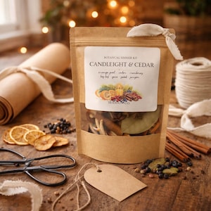 May include: A brown paper bag with a clear window, labeled "Candlelight & Cedar Botanical Simmer Kit." The bag contains dried botanicals. Dried orange slices, scissors, and a blank tag are on a wooden surface.