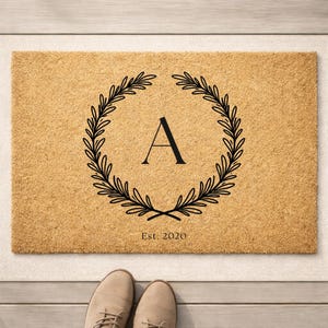 May include: A rectangular doormat with a natural tan color. A black floral wreath surrounds the letter "A" in the center. The text "Est. 2020" is below the letter. The doormat is on a light-colored surface.