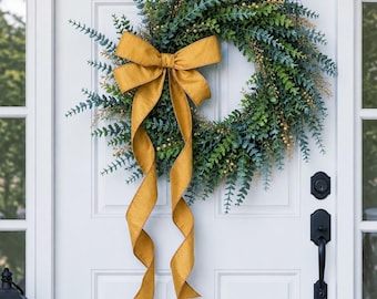 May include: A circular wreath composed of green eucalyptus leaves and small golden berries, featuring a large, mustard-yellow fabric bow tied at the top with two long, flowing ribbon tails hanging down the center of the foliage arrangement.