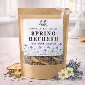 May include: A brown paper bag labeled "Stovetop Simmer Kit Spring Refresh" with a clear window showing dried herbs and fruit. The label includes the words "citrus, herbal, soft floral" and is decorated with blue flowers. A white ribbon is tied at the top.