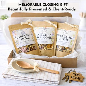 May include: A gift box with three pouches labeled "Home Sweet Home", "Kitchen Welcome", and "Welcome Home". A wooden spoon rests on a ceramic spoon rest, and a set of keys with a "New Home" tag are included.