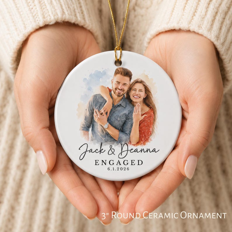 K&ouml;nnte beinhalten: Runder Keramikschmuckanh&auml;nger, ca. 7,6 cm Durchmesser, mit einem Aquarellbild eines Paares. Der Anh&auml;nger tr&auml;gt die Namen "Jack & Deanna", das Wort "ENGAGED" und das Datum "6.1.2026". Er hat eine goldene Aufh&auml;ngeschnur.