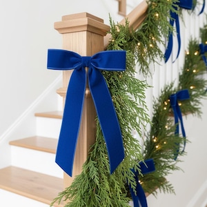May include: A staircase banister decorated with evergreen garland and vibrant blue velvet bows. The garland is adorned with small, warm white lights, creating a festive and inviting atmosphere. The bows are tied at intervals along the banister.
