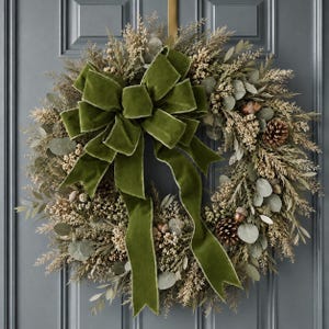 May include: A circular wreath featuring dried foliage, eucalyptus leaves, pinecones, and small acorns. A large, prominent olive green velvet ribbon with long, flowing tails is tied in a bow at the top center, creating a decorative focal point.