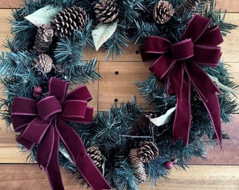 May include: A festive Christmas wreath with faux evergreen branches, pinecones, and burgundy velvet bows. The wreath is decorated with small red berries and light green leaves, creating a classic holiday decoration.