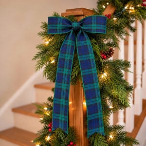 May include: A decorative bow made of blue and green plaid fabric with two long, hanging tails. The bow is attached to a green garland featuring small white string lights and red berries, wrapped around a wooden staircase banister post.