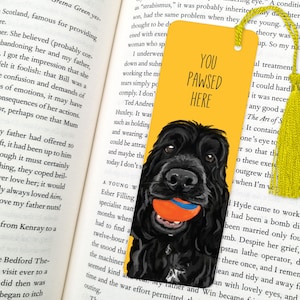 May include: Yellow bookmark featuring a black dog illustration holding an orange ball. The bookmark has the text "You Pawsed Here" in black.