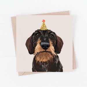 May include: A birthday card illustration of a dachshund wearing a gold glitter party hat with an orange pom-pom. The dog has brown and black fur and a tan muzzle. The card is on a light tan background with a matching envelope.
