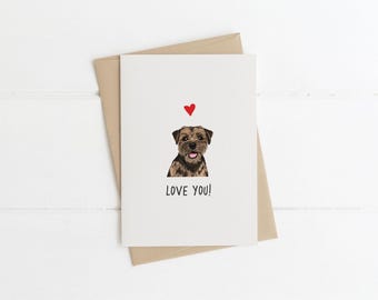 May include: A white greeting card featuring a brown and black dog illustration with a small red heart above its head and the text LOVE YOU! printed in black below the dog. The card is placed on top of a light brown envelope.