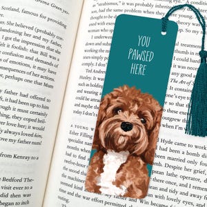 May include: Teal bookmark featuring a brown and white dog illustration and the text "YOU PAWSED HERE". It has a teal tassel and is placed in an open book. The bookmark is a reading accessory.