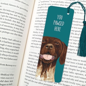 May include: Teal blue bookmark with a brown and white dog illustration. The bookmark has the text "You Pawsed Here" in white.