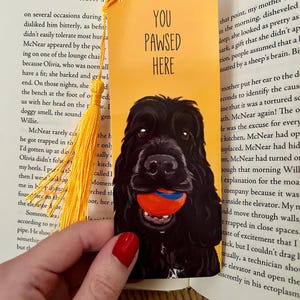 Könnte beinhalten: Ein gelbes, rechteckiges Lesezeichen mit dem Text YOU PAWSED HERE und einer Illustration eines schwarzen Hundes, der einen roten und blauen Ball im Maul hält. Das Lesezeichen hat eine gelbe Quaste, die durch ein Loch oben befestigt ist.