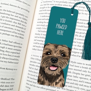 May include: Teal blue bookmark featuring an illustration of a brown and white dog. The bookmark has the text "You Pawsed Here" in white lettering.