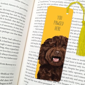 May include: A yellow bookmark with a watercolor illustration of a brown dog and the text "YOU PAWSED HERE." The bookmark has a yellow tassel and is placed in an open book.