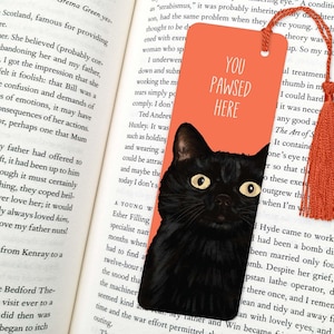 Peut inclure: Un chat noir aux yeux verts se cache derri&egrave;re un marque-page orange vif. Le marque-page porte le texte "You Pawsed Here" en blanc.