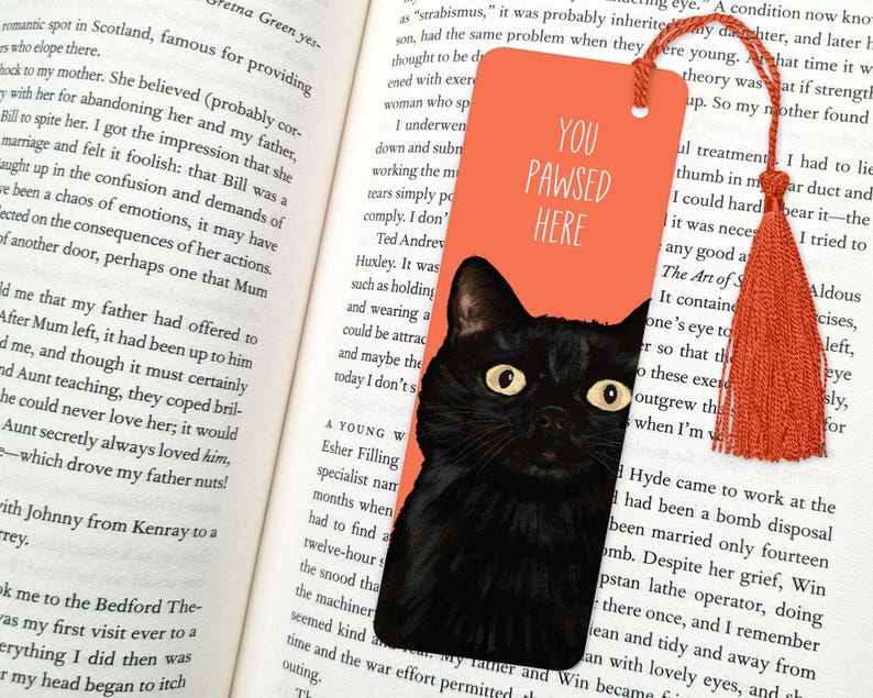 Peut inclure: Un chat noir aux yeux verts se cache derri&egrave;re un marque-page orange vif. Le marque-page porte le texte "You Pawsed Here" en blanc.