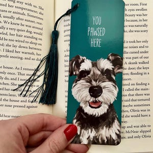 Peut inclure: Un marque-page rectangulaire sarcelle orné d'une illustration peinte d'un chien Schnauzer noir et blanc avec le texte YOU PAWSED HERE en lettres blanches. Un pompon sarcelle foncé est attaché au coin supérieur gauche par un cordon.