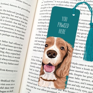 May include: Teal blue bookmark with a painted illustration of a brown and white dog with a pink tongue sticking out. The bookmark has the text "You Pawsed Here" in white.