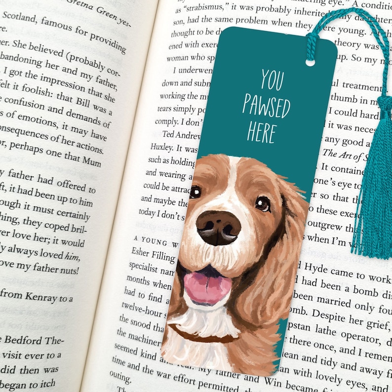 May include: Teal blue bookmark with a painted illustration of a brown and white dog with a pink tongue sticking out. The bookmark has the text "You Pawsed Here" in white.