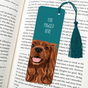 May include: Teal blue bookmark with a cartoon illustration of a brown and white dog with a pink tongue sticking out. The bookmark has the text "You Pawsed Here" in white.
