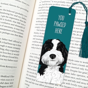 May include: Teal bookmark with a black and white dog illustration and a teal tassel. The bookmark reads "YOU PAWSED HERE" in white text. The bookmark is placed in an open book.