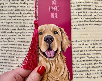 May include: A rectangular bookmark with a magenta background featuring a golden retriever illustration and the white text YOU PAWSED HERE. The bookmark has a hole at the top with a bright pink tassel attached, resting against an open book page.