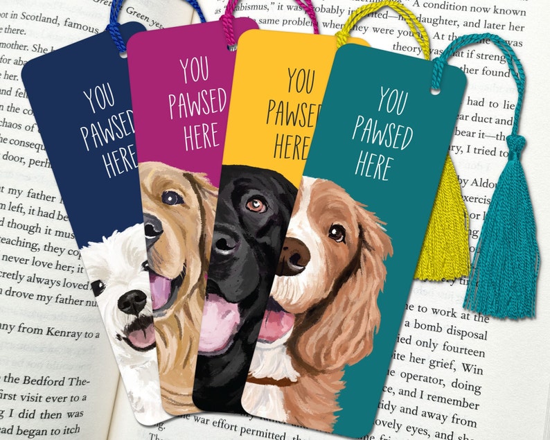 May include: Four colorful bookmarks with different dog breeds. Each bookmark features the text "You Pawsed Here" in white lettering. The bookmarks are teal, yellow, pink, and navy blue.
