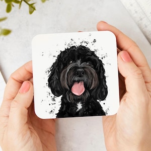May include: A square coaster featuring a watercolour-style illustration of a black dog with a white chest patch, floppy ears, and a pink tongue hanging out. The background is white with black paint splatters surrounding the dog's head.