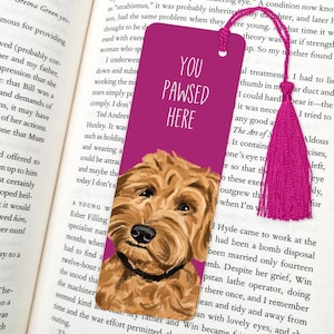 May include: A magenta bookmark with a watercolor illustration of a golden doodle dog. The text "YOU PAWSED HERE" is printed in white. A matching magenta tassel hangs from the top.