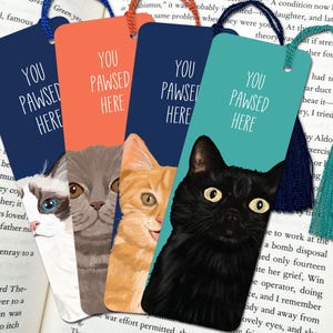 May include: Four rectangular bookmarks featuring illustrations of a white and grey cat, a grey cat, an orange tabby cat, and a black cat. Each bookmark displays the text YOU PAWSED HERE and includes a colored string tassel at the top.