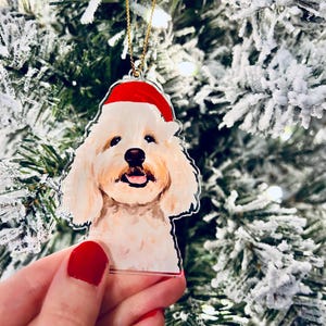May include: A clear acrylic Christmas ornament featuring a smiling dog wearing a red Santa hat. The dog has cream-colored fur and a black nose. The ornament hangs from a gold-colored chain. The background is a blurred Christmas tree.