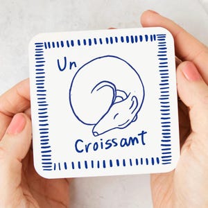 May include: Square white coaster with a blue line drawing of a curled-up dog and the words "Un Croissant" in blue. The coaster has a blue dotted border and is held by two hands.