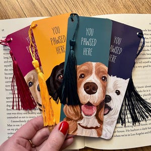 以下が含まれることがあります： 「YOU PAWSED HERE」という文字と犬の顔のイラストが描かれた4枚の長方形のしおり。赤、黄、ダークティール、黒のタッセルが付いています。しおりの地色はマゼンタ、オレンジ、ティール、ダークパープルです。