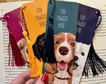 May include: Four rectangular bookmarks featuring illustrations of dog faces and the text "YOU PAWSED HERE". Each bookmark has a colored tassel attached: red, yellow, dark teal, and black. The bookmarks are in shades of magenta, orange, teal, and dark purple.