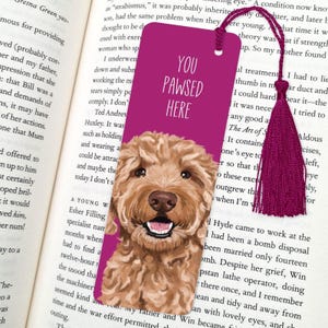 May include: A pink bookmark featuring a doodle of a light brown dog with a white chest and a pink nose. The bookmark has the text "You Pawsed Here" in white letters.