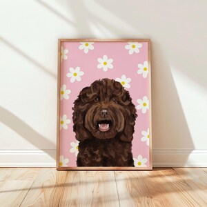 May include: A framed rectangular wall art print featuring a digital illustration of a dark brown curly-haired dog with an open mouth. The background is a solid light pink colour decorated with a repeating pattern of white daisies with yellow centres.