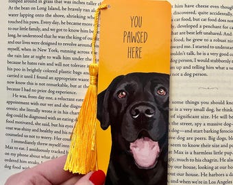 May include: A rectangular yellow bookmark featuring a black Labrador dog illustration and the text "YOU PAWSED HERE" in black lettering. The bookmark includes a bright yellow tassel attached to the top left corner with a small metal eyelet.