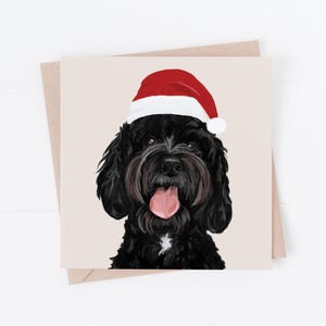 May include: A square Christmas card showing a black dog wearing a red and white Santa hat. The dog has its tongue out. The card is on a beige background with a brown envelope.
