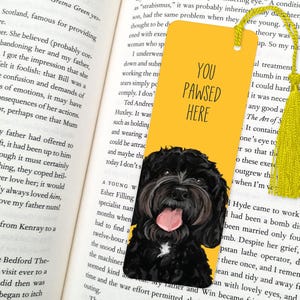 May include: A yellow bookmark with a black and white illustration of a dog's head. The bookmark has the text "You Pawsed Here" on it.