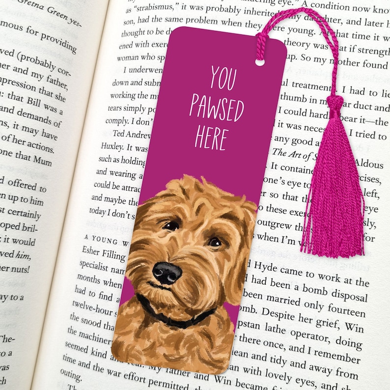 May include: A pink bookmark with a cute illustration of a brown and white dog. The bookmark has the text "You Pawsed Here" in white letters.