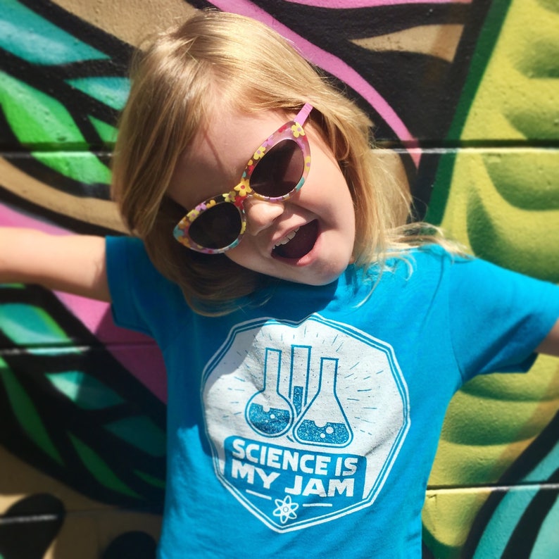 May include: A young girl wearing a blue t-shirt with the text "Science is my jam" and a white graphic of beakers. She is wearing pink and floral patterned sunglasses and has a bright, excited expression.
