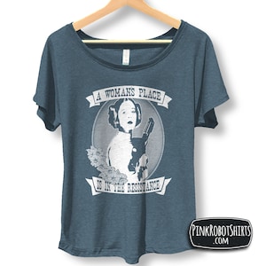 Könnte beinhalten: Ein blaues T-Shirt mit einem weißen Aufdruck von Prinzessin Leia, die einen Blaster hält. Der Aufdruck enthält den Text "A Woman's Place Is In The Resistance".