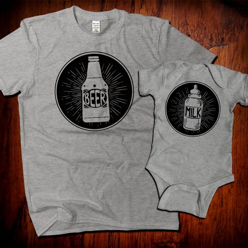 May include: A gray t-shirt with a black circle design featuring a beer bottle with the word "BEER" on it. A gray baby onesie with a black circle design featuring a baby bottle with the word "MILK" on it.