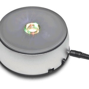 May include: A silver rotating display stand with a clear glass top and a small LED light in the center. The light is on and is emitting a green and blue glow. The stand is plugged in with a black cord.
