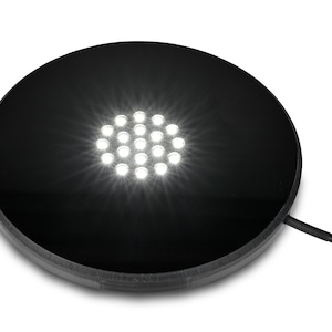 May include: A black, round LED display base with a cluster of bright white lights in the center. The base has a smooth, reflective surface and a textured edge. A black power cord extends from the side.